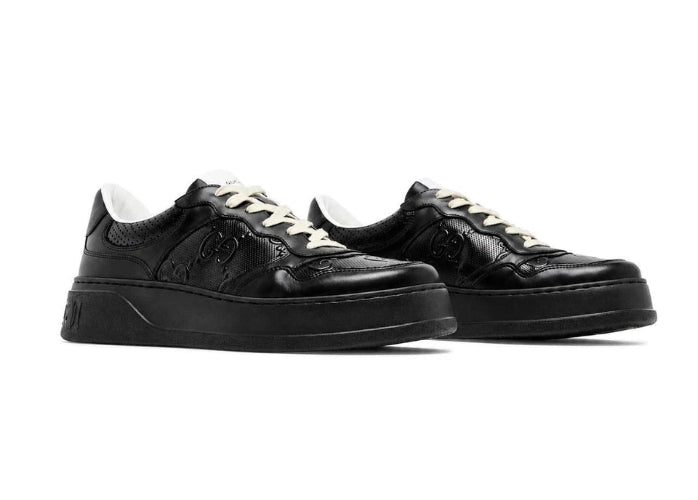 A pair of black leather low-top lace-up sneakers with black soles, shown from a side profile view against a plain white background.