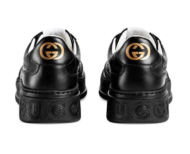 A pair of black leather low-top sneakers shown from the back, featuring a prominent interlocking GG logo in gold-tone metal on the heel counters.