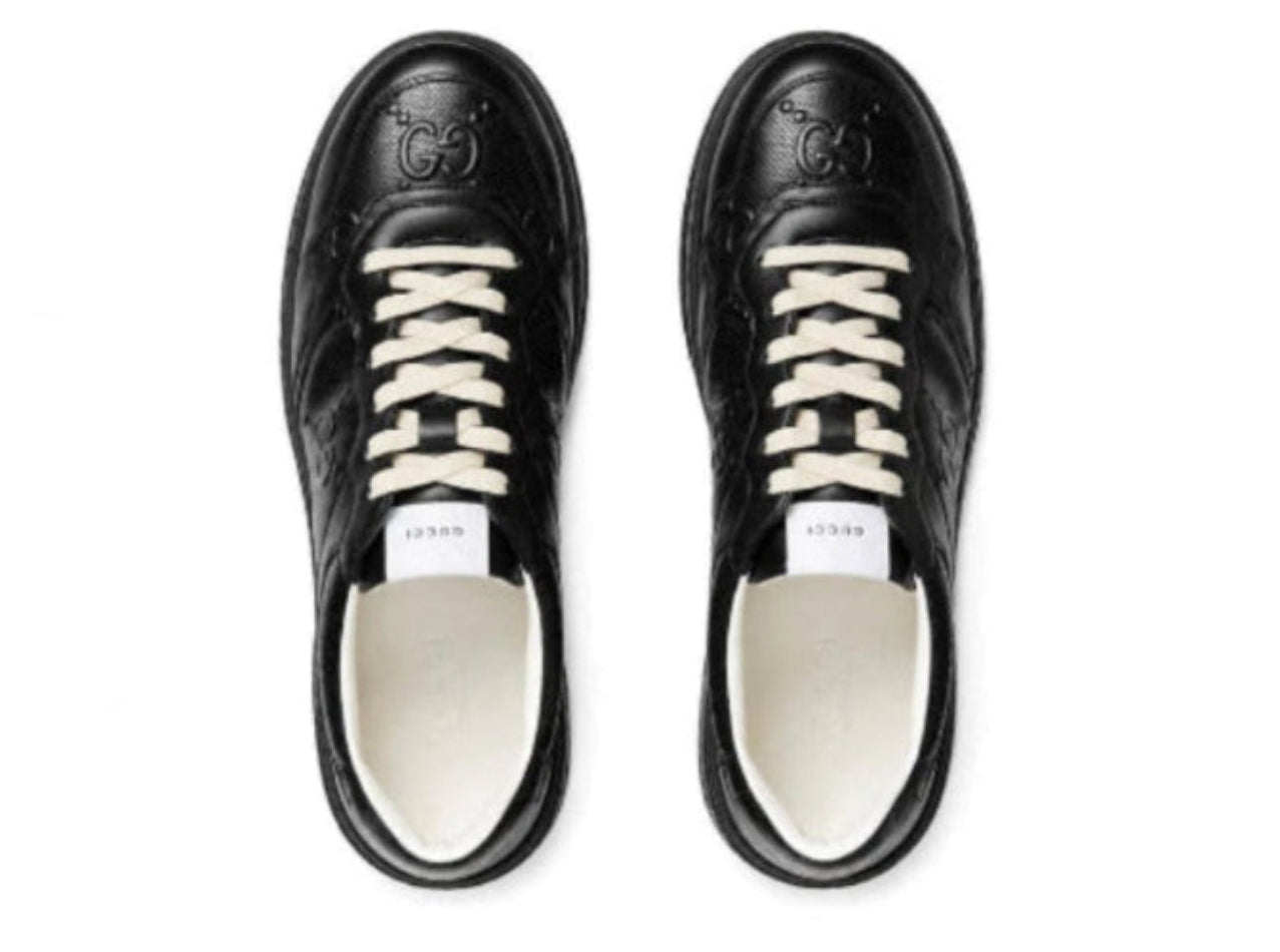 A pair of black leather Gucci sneakers shown from a top-down view on a white background. The shoes feature embossed GG logos on the toe, white laces, and a white Gucci label on the tongue.