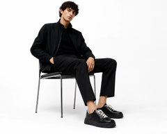 A model wearing a black long-sleeve top, black trousers, and black shoes sits on a black metal chair against a plain white background.