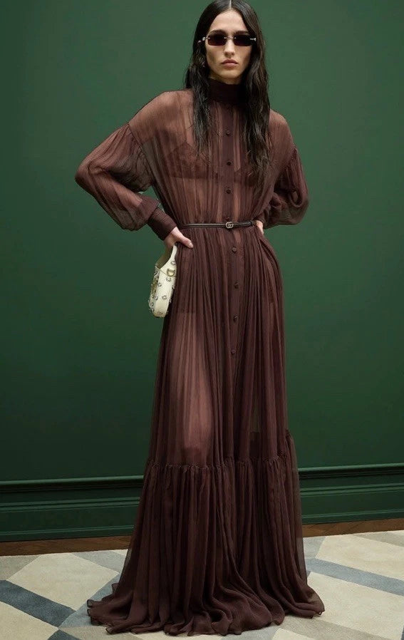 A model wearing a long-sleeved, high-neck, button-front maxi dress in a deep brown or burgundy chiffon-like fabric, styled with a matching belt and a small white handbag, set against a dark green wall.
