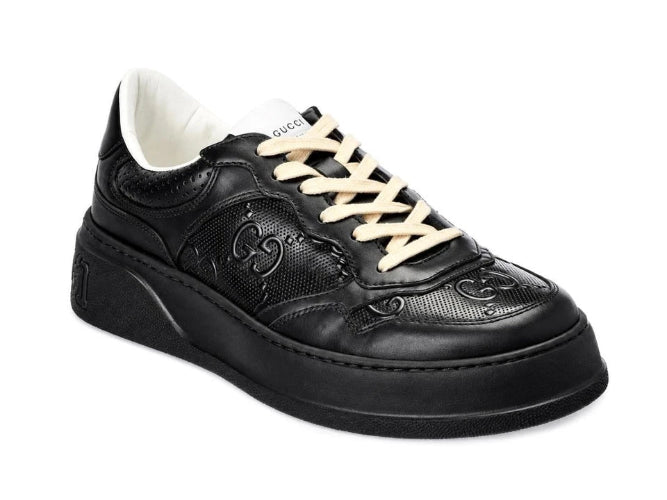 A black leather low-top sneaker with black laces and a thick black sole, shown against a plain white background.
