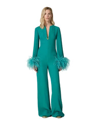 A woman modeling a teal green long-sleeve jumpsuit with feathered cuffs and a split neckline, photographed against a plain light beige studio background.