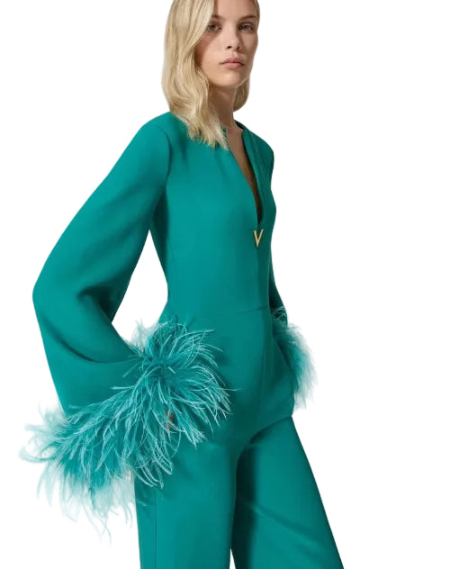 A model wearing a teal green jumpsuit with dramatic bell sleeves featuring turquoise feather trim at the cuffs, photographed against a solid off-white background.