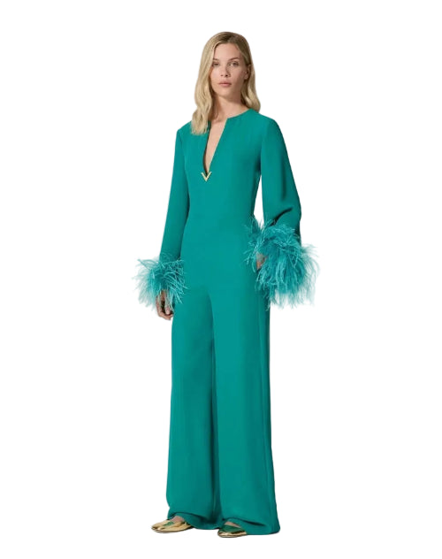 A woman modeling a teal green wide-leg jumpsuit with long sleeves, a deep V-neckline, and matching feathered cuffs, set against a plain light beige background.