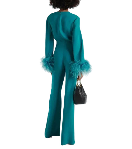 A model is shown from the back wearing a teal green long-sleeve jumpsuit with feathered cuffs, holding a small black handbag against a plain white studio background.