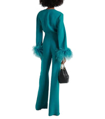 A model is shown from the back wearing a teal green long-sleeve jumpsuit with feathered cuffs, holding a small black handbag against a plain white studio background.