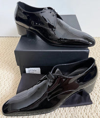 A pair of men's black patent leather dress shoes displayed on top of their black shoebox against a light beige textured background.