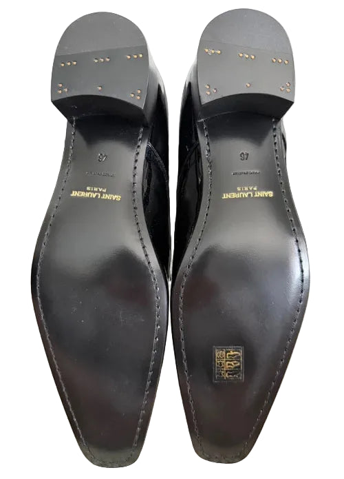 A pair of black leather dress shoes shown from the sole side, positioned in front of their black Saint Laurent box on a beige carpet. The soles feature gold lettering reading 'SAINT LAURENT PARIS' and a gold square logo patch.