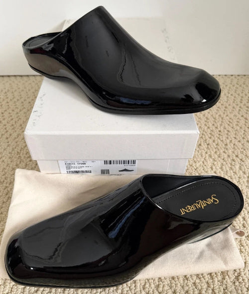 A pair of black patent leather mule flats with a low block heel, displayed on a white shoebox and white fabric surface.