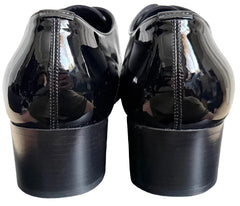 A pair of black patent leather oxford shoes shown from the back, featuring a glossy finish and stacked block heels, set against a neutral gray background.