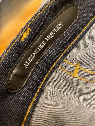 Close-up of a pair of dark blue denim jeans showing the inner waistband label which reads 'ALEXANDER MCQUEEN' in white text, with yellow contrast stitching.