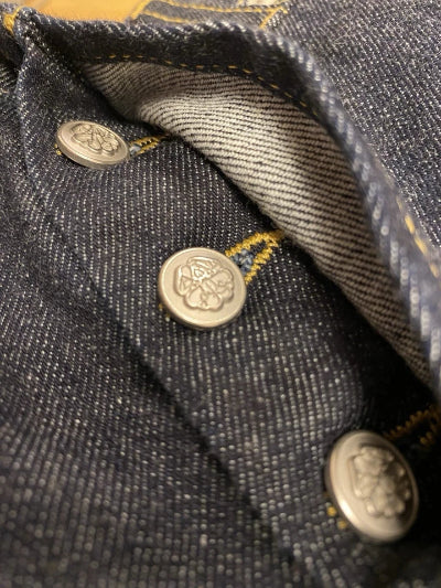 Close-up of dark indigo denim fabric with visible silver-tone buttons and yellow contrast stitching.
