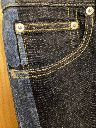 Close-up of a denim jacket or denim fabric showing a dark indigo front pocket with contrast yellow stitching, a lighter blue pocket panel, and silver metal rivets, photographed against a light wood surface.