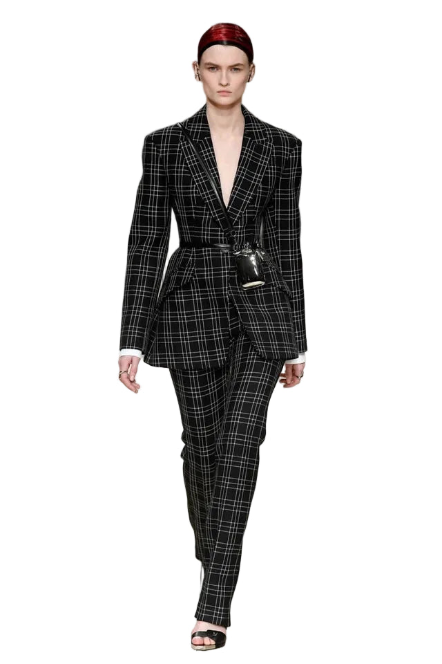 A model walks down a runway wearing a black and white plaid blazer and matching trousers, styled with a small black crossbody bag and a red headband.
