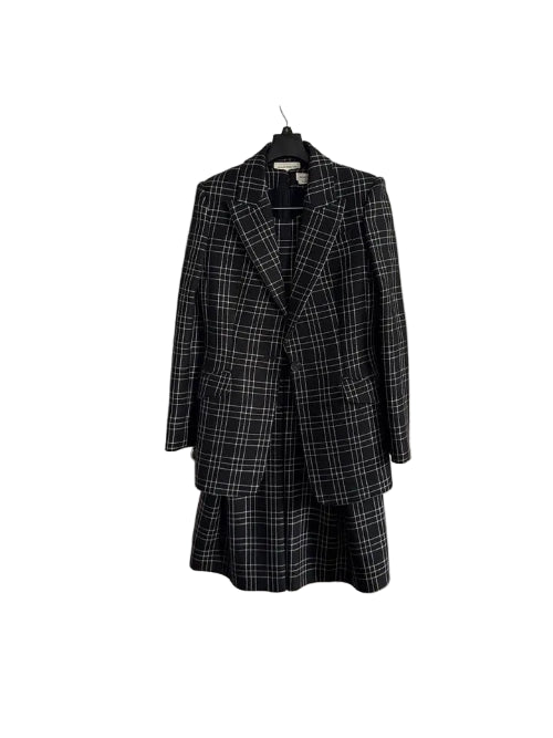A black and white plaid blazer and matching skirt set hanging on a black hanger against a white paneled door.