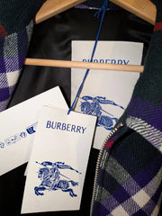 A close-up of a Burberry garment on a wooden hanger, showing a plaid pattern in dark green, purple, white, and black, along with white paper tags printed with the blue Burberry logo and equestrian emblem.