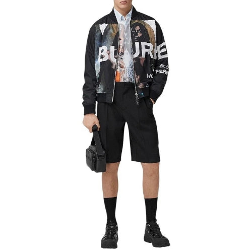 A model wearing a black bomber jacket with a large graphic print on the front, styled with black shorts, black socks, and black boots, holding a small black bag against a plain white background.