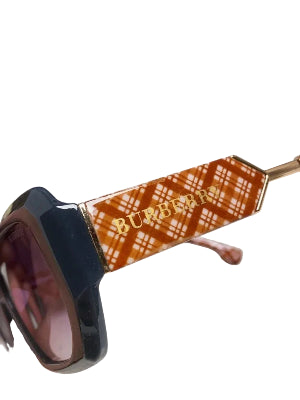 Close-up of a pair of sunglasses with a glossy black frame and a brown and white plaid acetate temple arm featuring gold 'Burberrys' lettering, held by a hand against a blue patterned background.