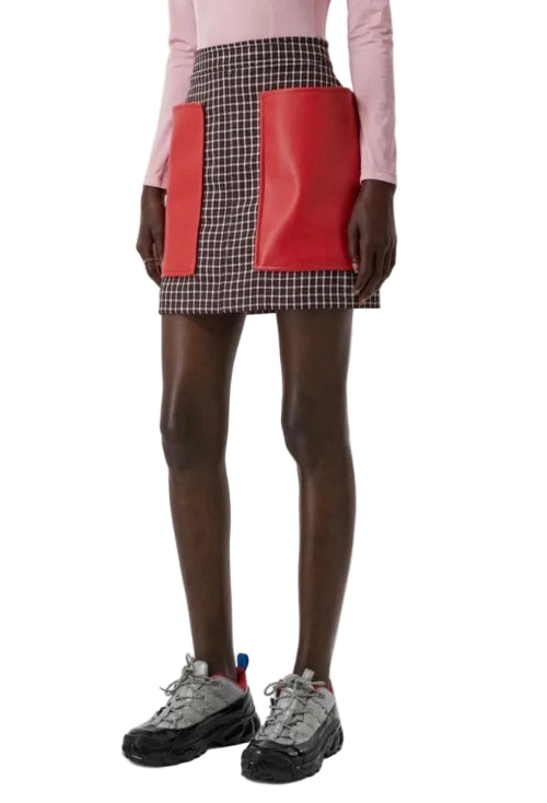 A model wearing a high-waisted mini skirt with a brown and white gingham pattern and two large red leather front pockets, styled with a pink long-sleeve top and chunky black sneakers.