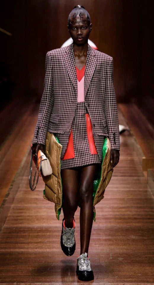 A model walks down a runway wearing a gray blazer with a colorful striped shirt and a patterned green and gold handbag.