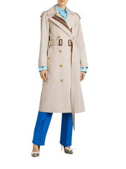 A model wearing a long beige trench coat with brown buttons and striped lapels, styled with bright blue trousers, a light blue shirt, and white pointed-toe heels against a light gray studio background.