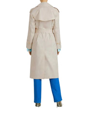 A model shown from the back wearing a light beige trench coat over bright blue trousers and metallic high heels, set against a plain light gray studio background.