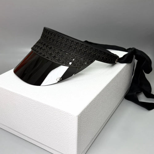 A black visor hat with a glossy visor, a textured patterned headband, and a black ribbon tie, displayed on a white textured box against a gray background.