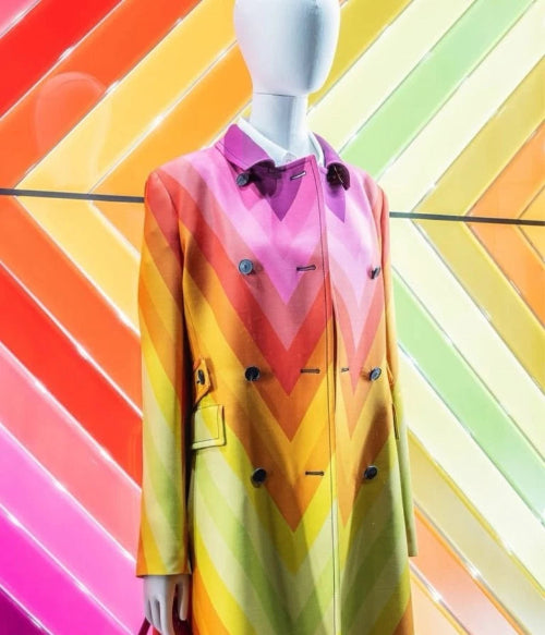 A colorful tie-dye long-sleeve shirt displayed on a white mannequin torso against a vibrant rainbow-striped background.