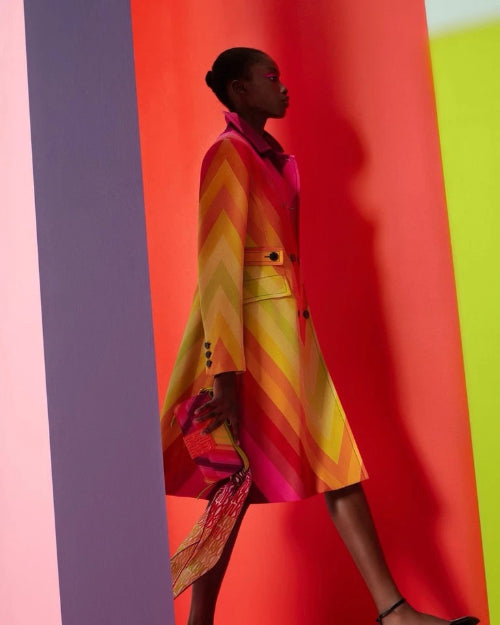 A model wearing a vibrant yellow and orange patterned dress walks against a bold, multi-colored background of vertical panels in pink, red, yellow, and purple.