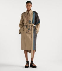 A model wearing a beige trench coat with blue and black color-blocked panels and floral embroidery, paired with black platform sandals, against a plain white studio background.