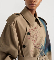 A close-up of a model wearing a beige double-breasted trench coat with floral embroidery on the front, paired with a blue and black patterned shirt underneath.