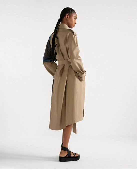 A model shown from the back wearing a mid-length beige trench coat with a belted waist, paired with black platform sandals and a dark backpack, against a plain light gray studio background.