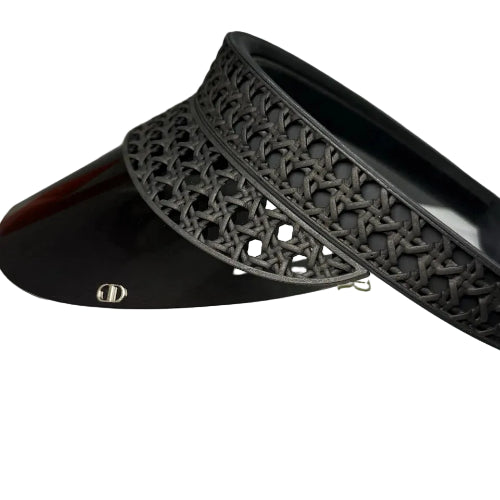 A close-up of a black visor hat with an intricate woven pattern on the brim and a glossy black shade, resting on a white textured surface.