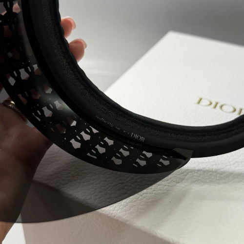 A close-up of a black Dior hat featuring an intricate geometric lattice cutout pattern on the brim, held by a hand over a white Dior-branded box.