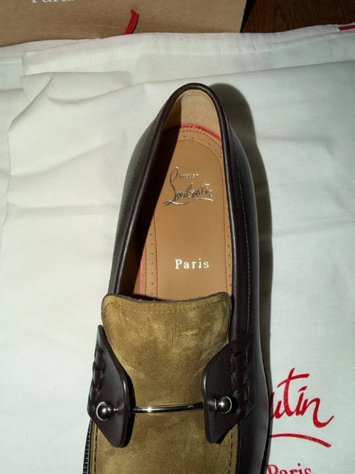 A Christian Louboutin Paris loafer in dark brown leather and olive green suede, displayed on its white dust bag with the brand's logo visible.