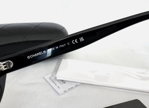 Close-up of a black Chanel sunglasses arm with white printed branding and text, resting on a white textured surface with a white dust bag and part of a black quilted case visible.