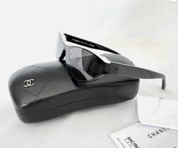 Chanel sunglasses with a black and white frame, resting on a black quilted Chanel case with a silver CC logo, alongside a white microfiber cloth and paper accessories on a white background.