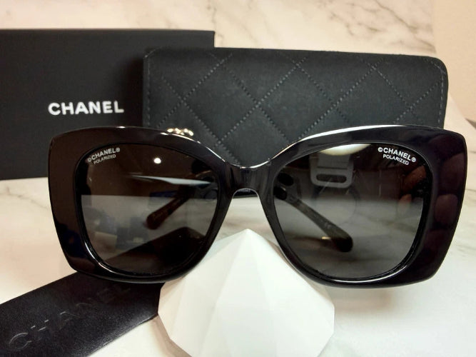CHANEL black cat-eye sunglasses displayed on a white stand in front of a black CHANEL box.
