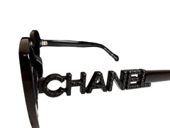 A pair of black Chanel sunglasses with a prominent silver-tone CHANEL logo on the frame, displayed next to its black branded box on a light marble surface.