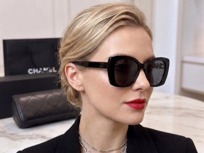 A woman wearing large black square sunglasses with a gold-tone frame, shown in a close-up lifestyle shot. A black box with gold lettering is visible in the background.