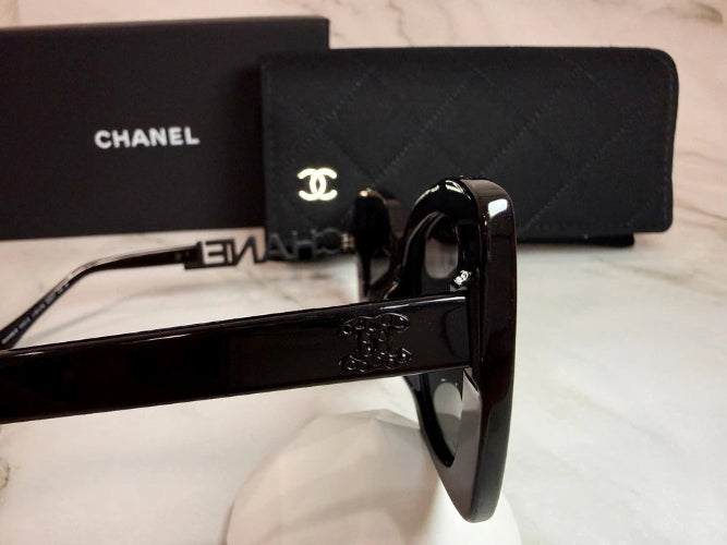 A pair of black Chanel sunglasses with the iconic interlocking CC logo on the temple, displayed next to its black presentation box on a light-colored surface.