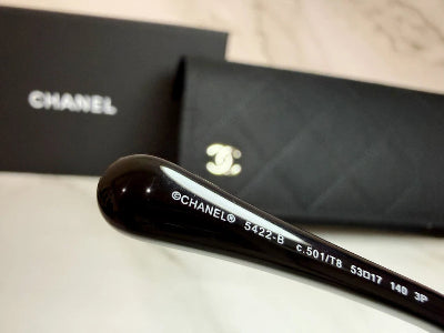 Close-up of a black Chanel eyeglass arm with white printed model text, shown in front of a black Chanel box and matching black quilted pouch on a light marble surface.