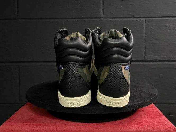 Black high-top sneakers with beige soles on a black platform against a dark brick wall.