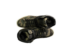 Camouflage high-top sneakers on a black stand against a dark brick wall.