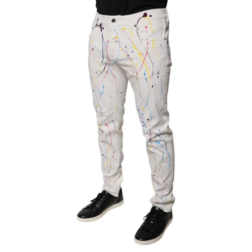 White slim-fit jeans with a colorful paint splatter print, shown on a model from the waist down wearing black sneakers against a white background.
