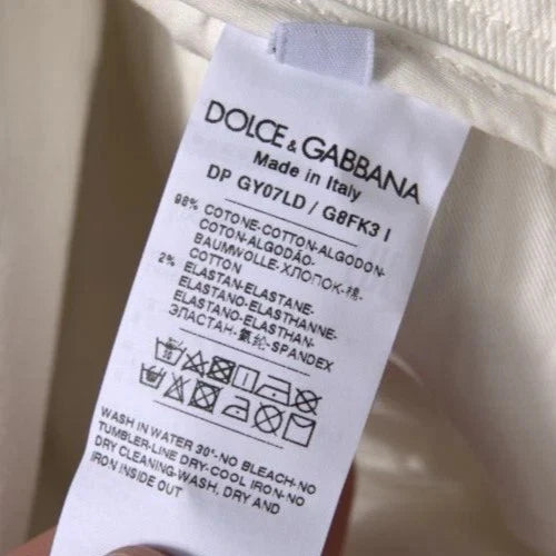 A close-up of a white fabric care label for Dolce & Gabbana clothing, showing product details and washing instructions, held against a light beige fabric background.