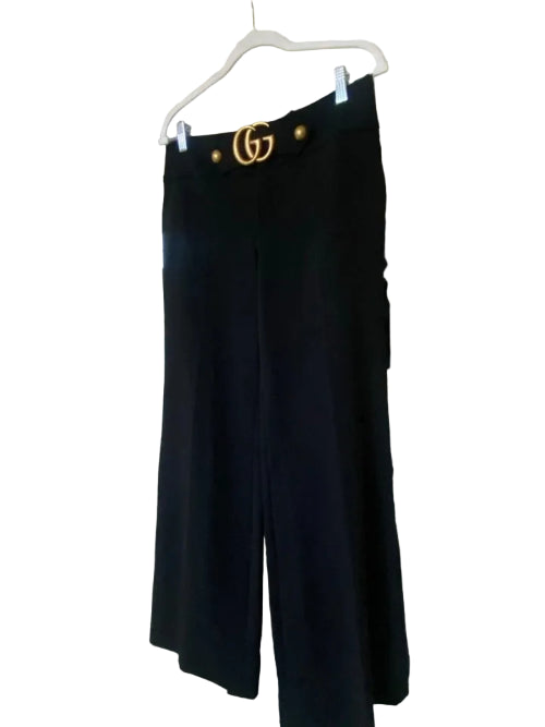 Black wide-leg pants hanging on a white hanger in front of a white door, with a gold logo on the waistband.