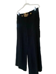 Black wide-leg pants hanging on a white hanger in front of a white door, with a gold logo on the waistband.