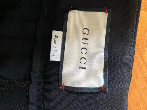 Close-up of a Gucci garment label sewn onto black fabric, with a smaller white 'Made in Italy' tag beside it. The main Gucci label is off-white with black lettering and red stitching, set against black material with a small area of wood surface visible at the right edge.